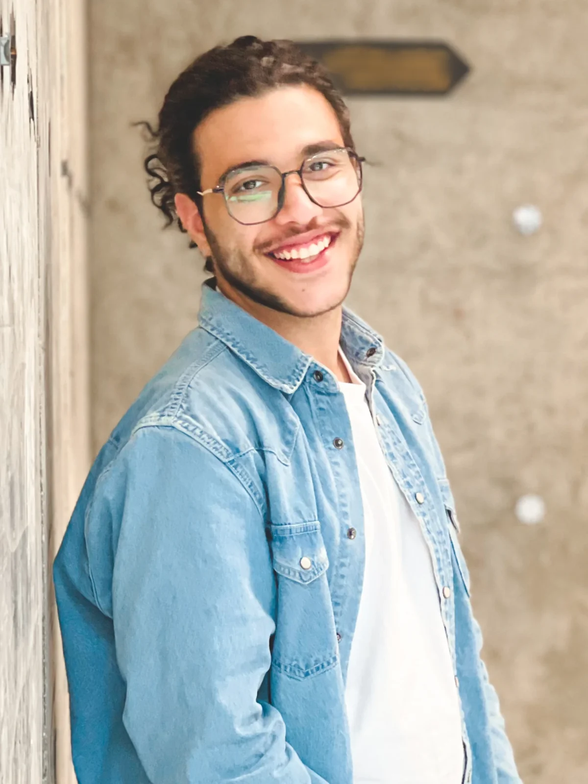 Danial Hadizadeh smiling, in glasses and a denim shirt, leaning on a wall